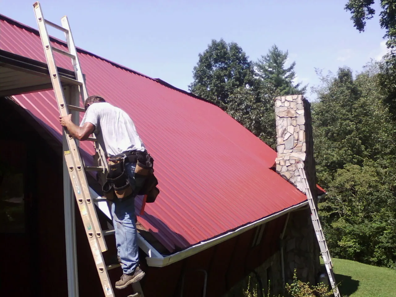 Roofer on a ladder beside a red metal roof and stone chimney — Soffit & Fascia Repair by Roofing Brothers in Gibbonsville